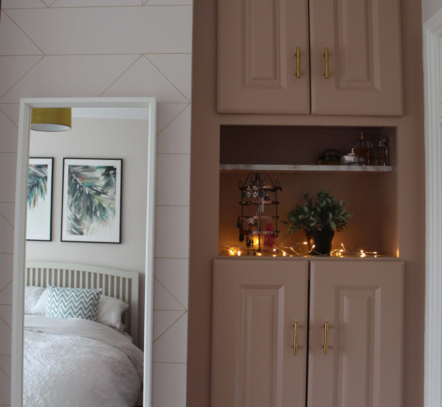 A mirror hanging on the left. On the right, a dusty pink wardrobe with a marble shelf holding fairy lights, a plant and jewellery. 