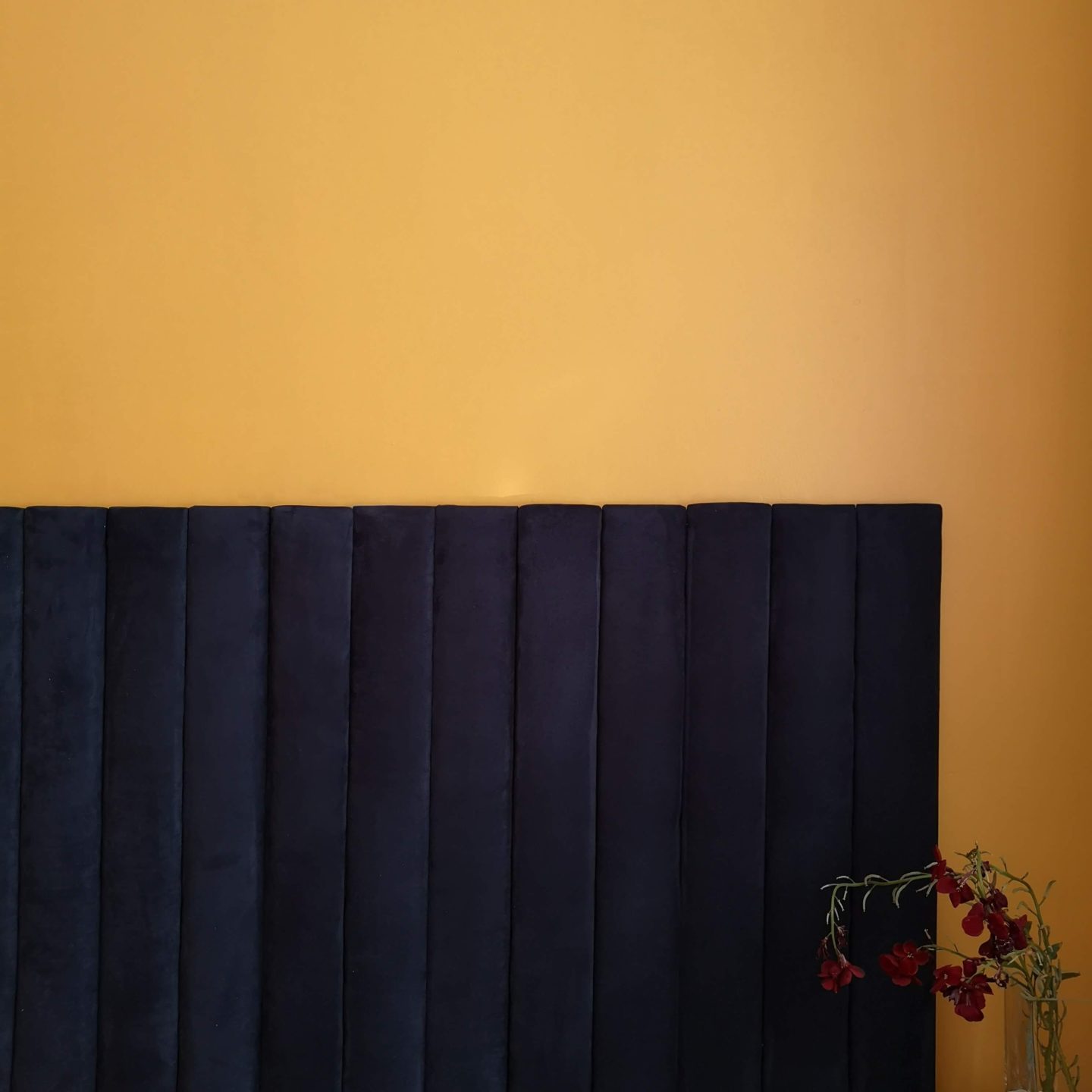Navy suede fabric headboard set against a mustard wall. Deep red wallflowers to the side.
