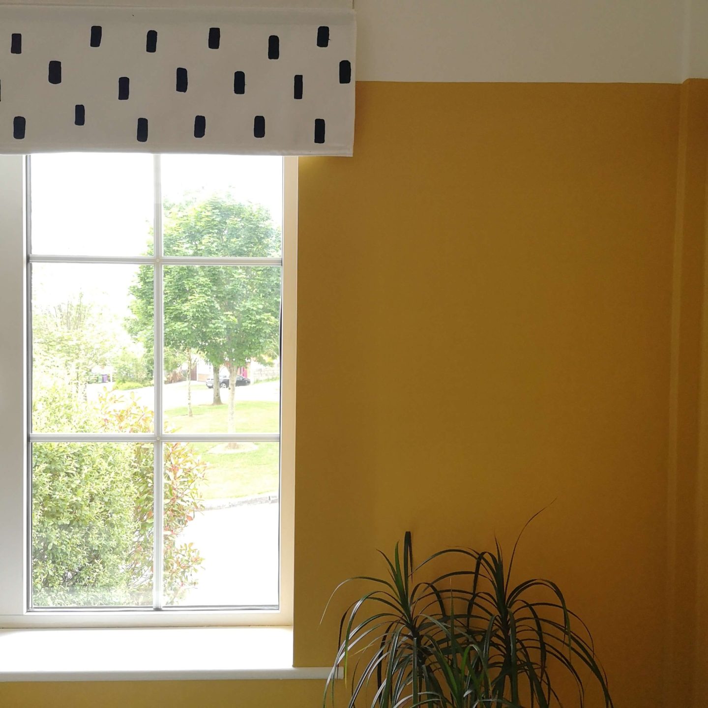 White blind with black rectangular pattern. Mustard yellow walls with green plant.