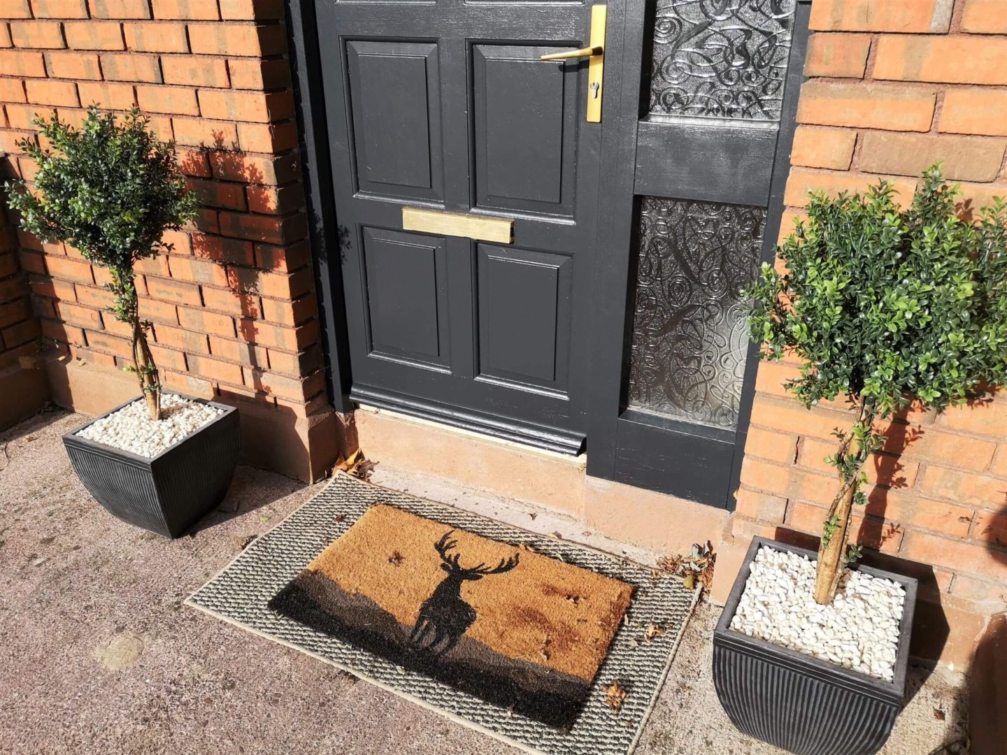 Grey front door, two black planters with small trees and two doormats layered on top of each other with a stag design on top.