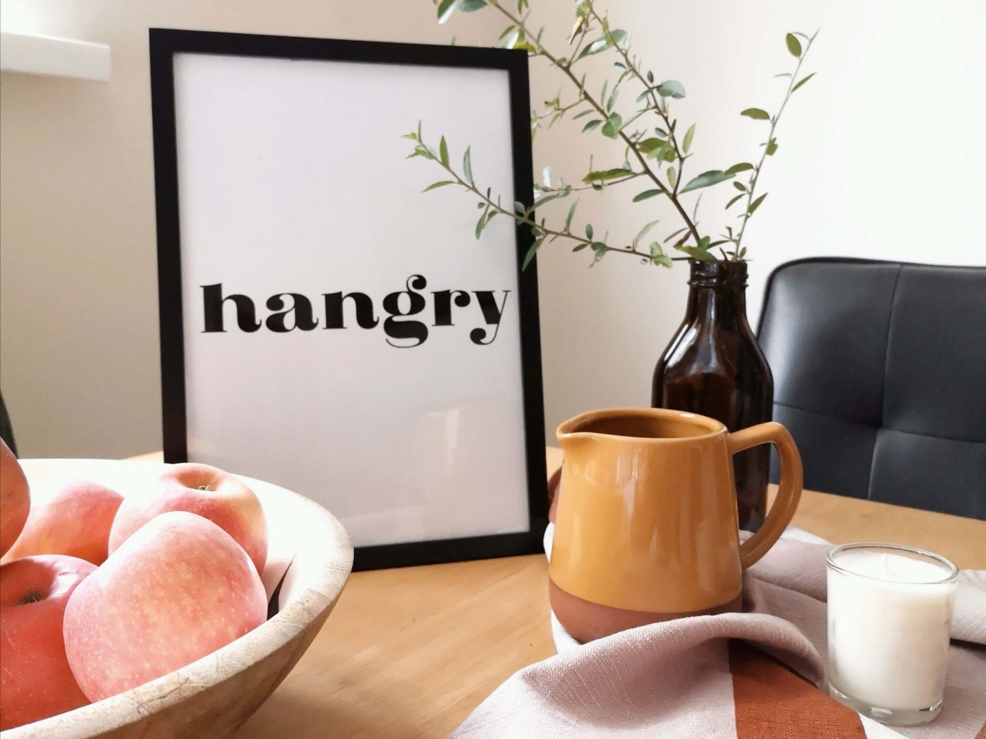 Black and white print of the word Hangry on a wooden dining table. Also present is a bowl of apples, mustard jug, greenery and an orange and pink tea towel.