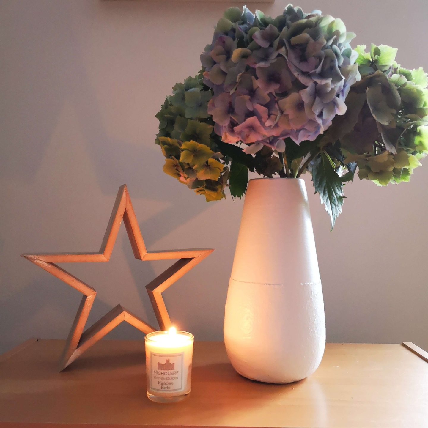 A white vase with blue and green hydrangeas standing next to a wooden start and small, white candle.