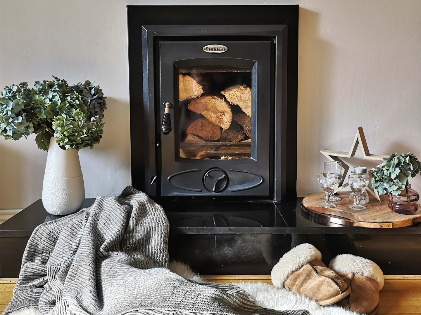 A matt black wood burning stove, unlit and filled with timber. It is surrounded by candles, a grey and white striped blanket, ugg slippers and hydrangeas in a white vase.