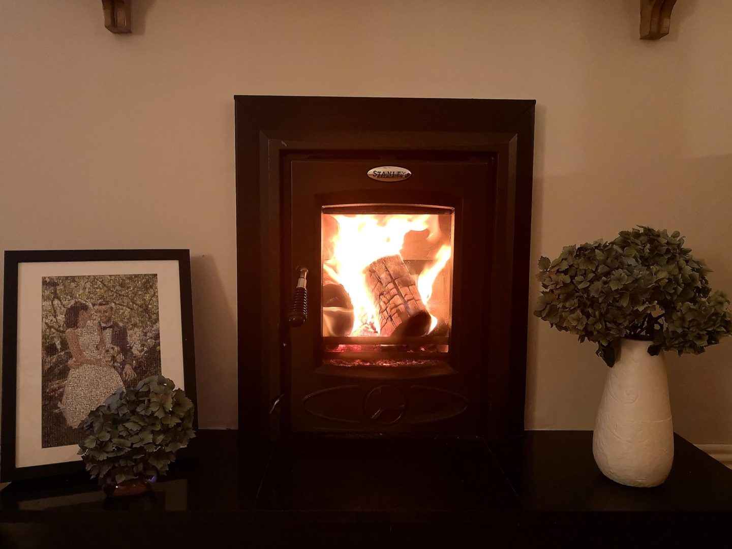 A black wood buring stove lighting at night. Light grey walls in the background and a black stone hearth in front. Surrounded by a wedding photograph and a white vase with hydrangeas. 