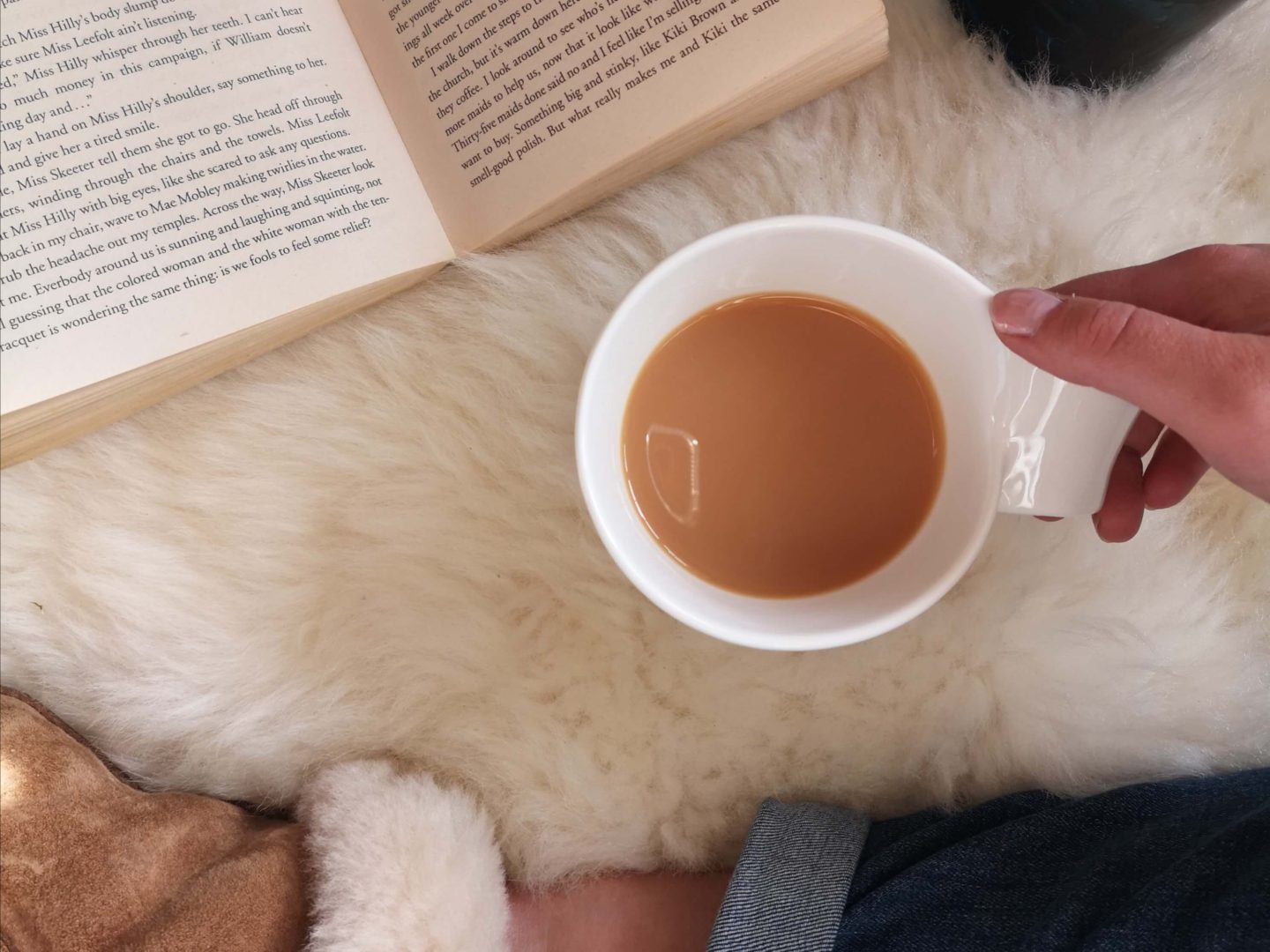 A flatlay of an open book, cup of tea and a person's leg wearing an ugg slipper. All on top of a sheepskin rug.