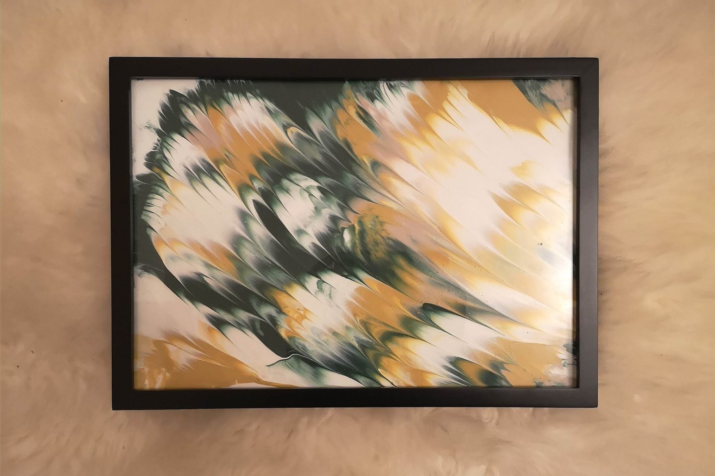 A colourful piece of art in a black frame, lying on a sheepskin rug. The colours in the artwork are white, green, yellow and pink.