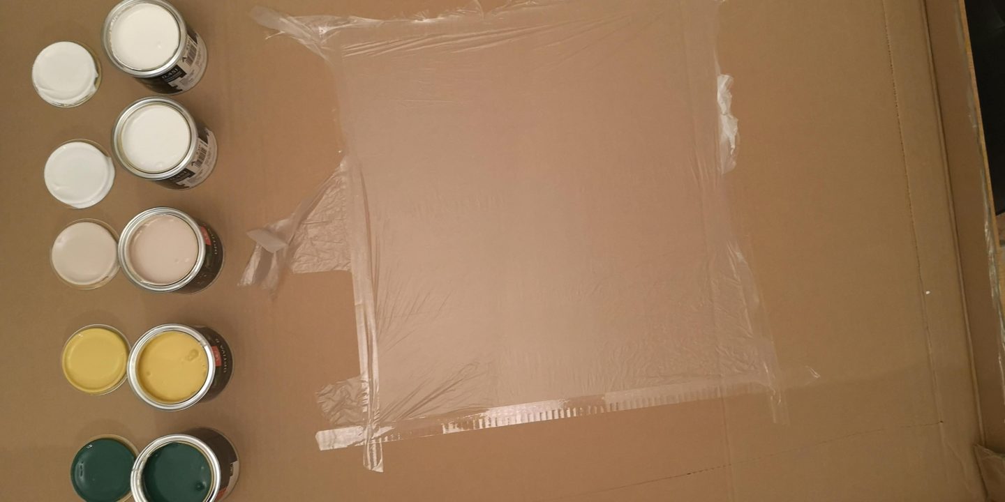 A large piece of cardboard with plastic taped to it and paint tins open nearby.