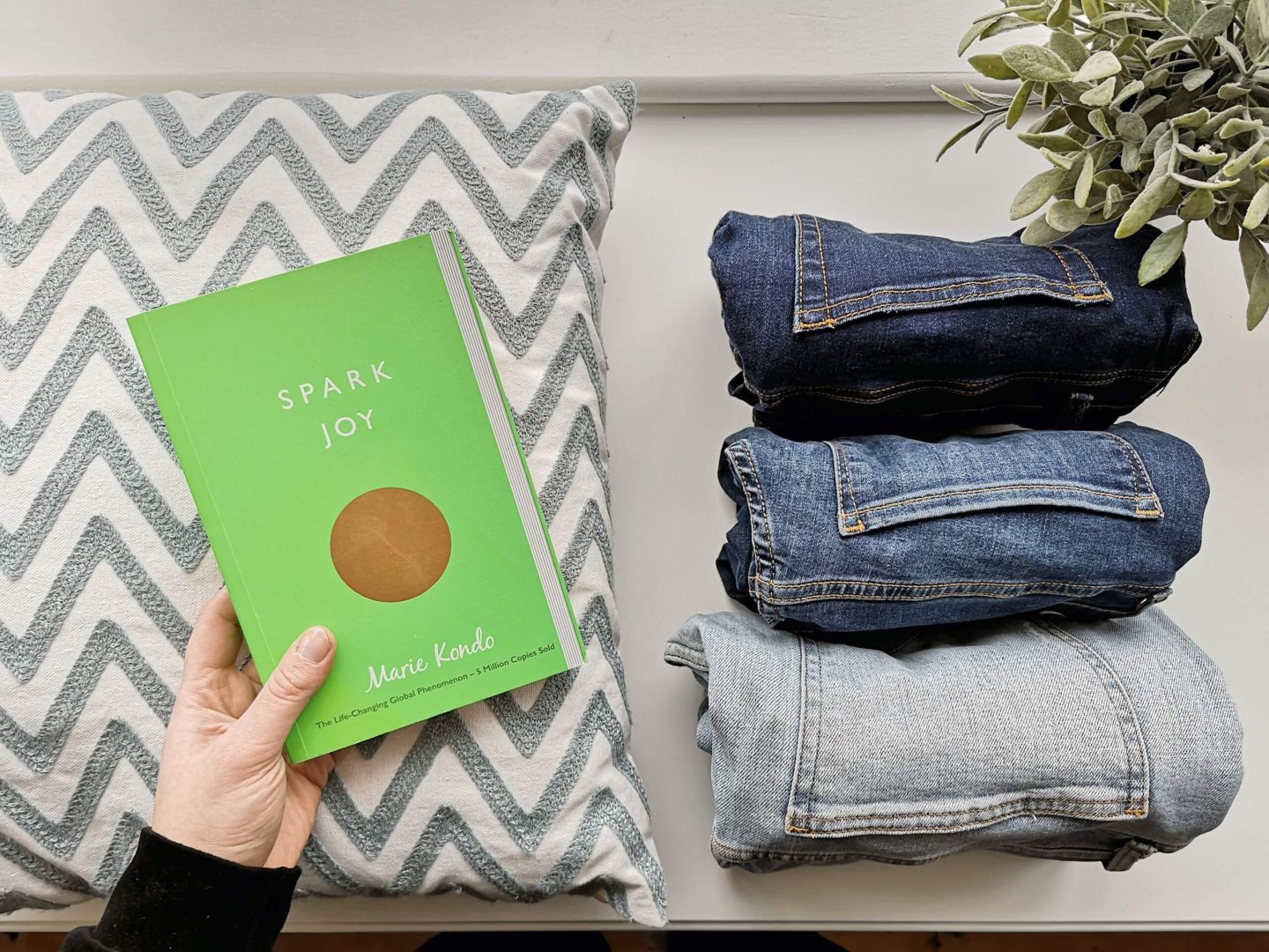 A flatlay of three pairs of jeans, a cushion, a plant and a copy of the Spark Joy book written by Marie Kondo. 