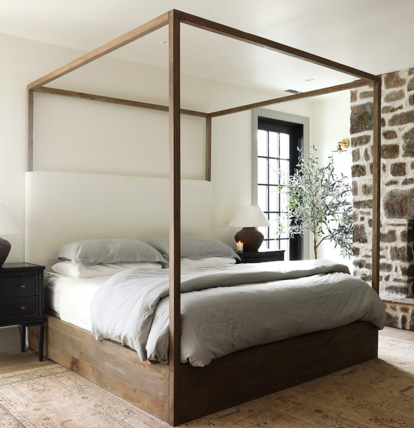 A white bedroom with a wooden canopy bed, a pale peach patterned rug, large clay lampshades and a stone fireplace.