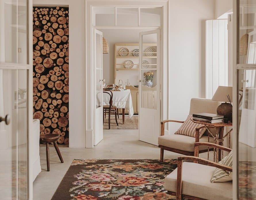 A living room with white walls, logs piled, white armchairs and a dark floral rug.