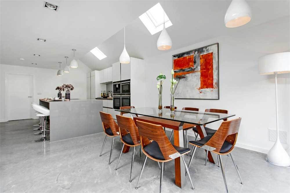 An open plan dining room and kitchen. Concrete floors and worktop with a glass dining table and dark wood dining chairs.