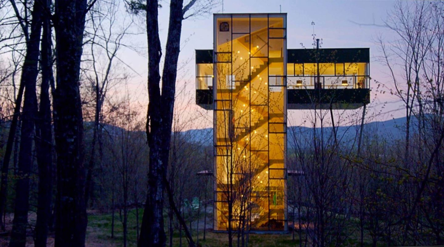 An unusual house shaped like the capital letter T. Picture taken at night and the central column are stairs up to a the main living area overlooking the trees around.