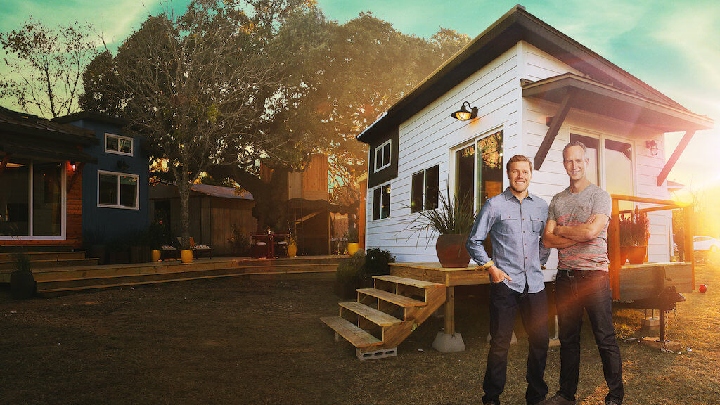 Two men standing outside a tiny house. The house has white wooden cladding with black trim. There are wooden steps leading up to the glass front doors.