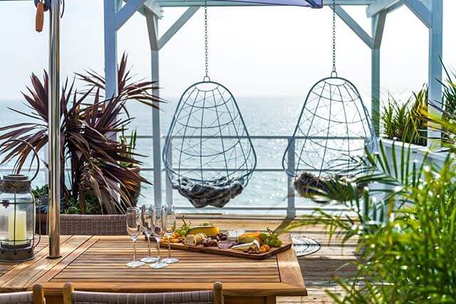 Two hanging egg chairs on a balcony overlooking the ocean. There are lots of plants on the balcony along with a dining table with a cheeseboard.