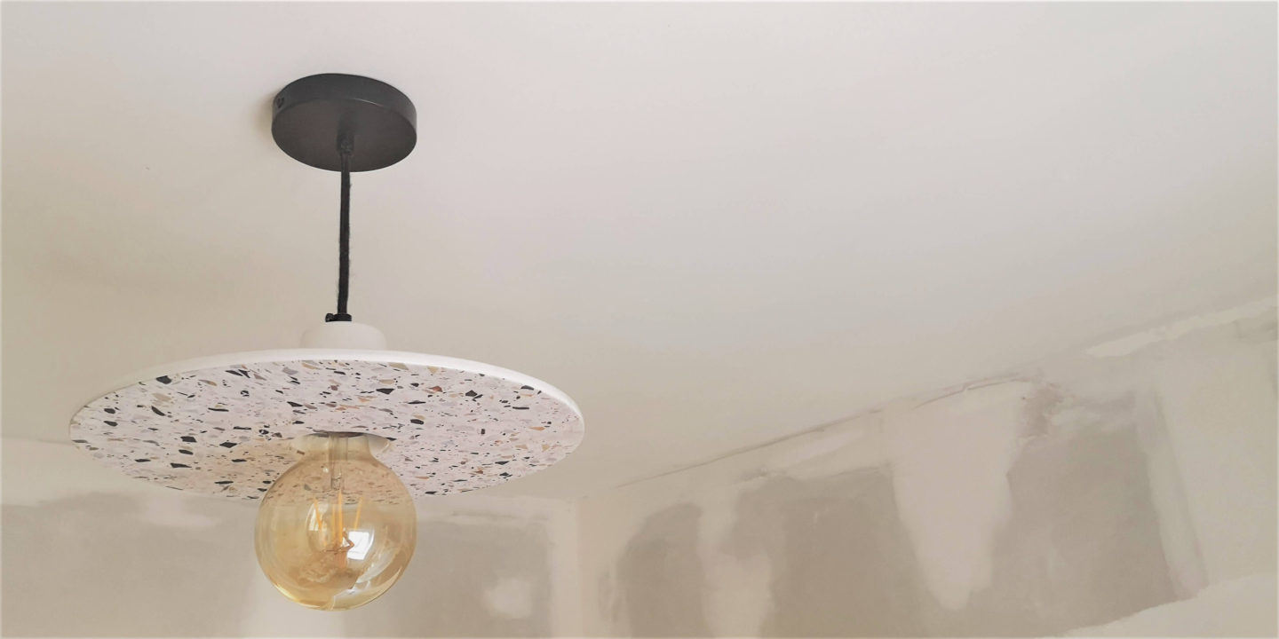 A plate shaped light with a terrazzo pattern and black cord hanging on an unpainted ceiling.