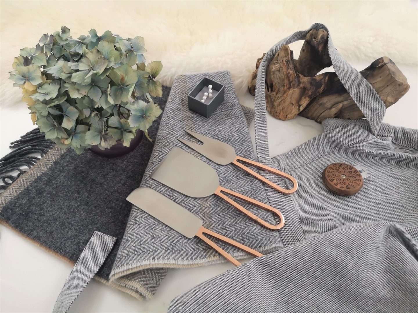 A flatlay showing a grey scarf, earrings, copper cheese knoves and a grey apron.