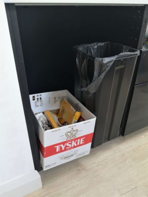 A black kitchen base cabinet without a door yet. A cardboard box is in there being used as a bin.