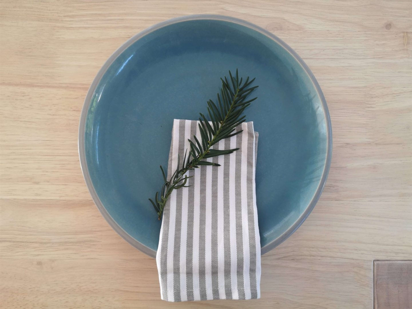 A blue dinner plate with a striped grey and white napkin on a light oak dining table.