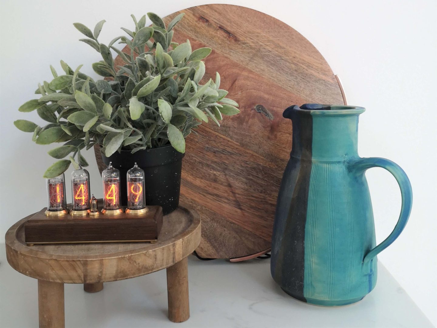 A neon tube clock on a wooden stand. A  round wooden board behind with with a blue jug and faux greenery.