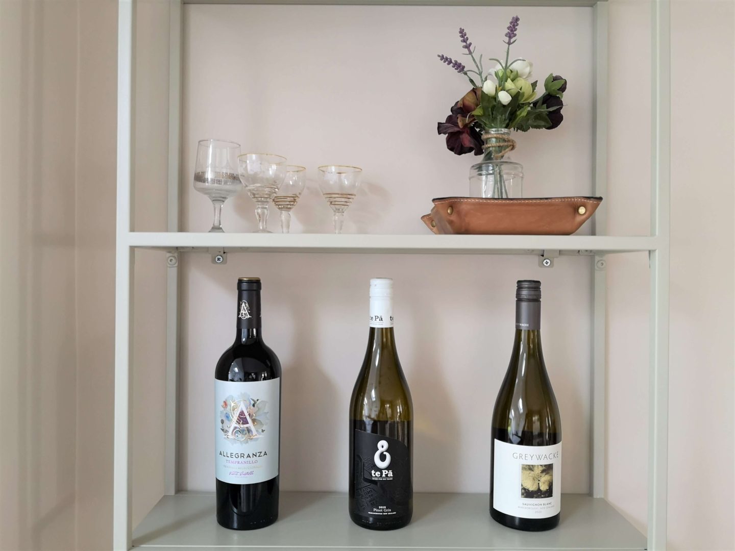 A close up of metal shelving, showing vintage glasses, flowers and three bottles of wine.