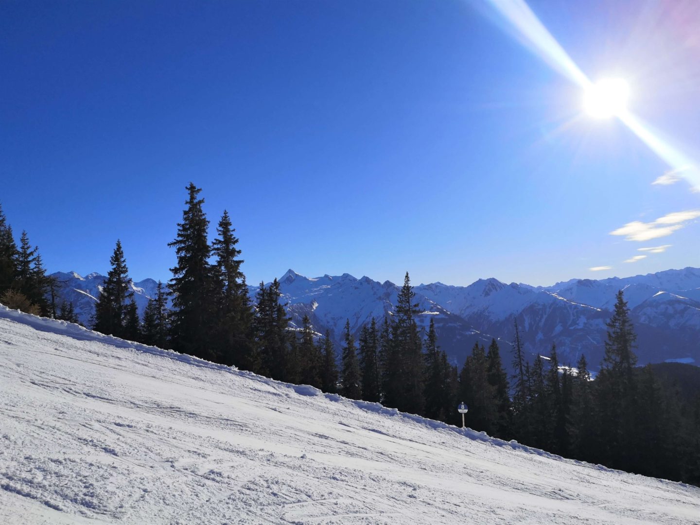 A ski slope with trees and mountain peaks in the background. A clear blue sky can be seen with the sun shining. 