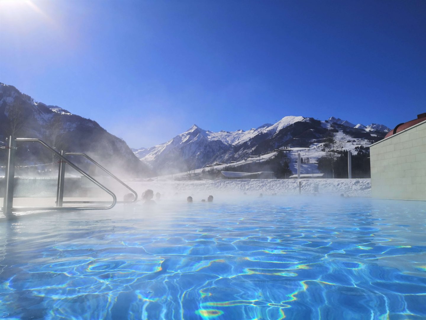 An outdoor pool, with steam rising. In the background are snowcapped mountains and a ski slope.