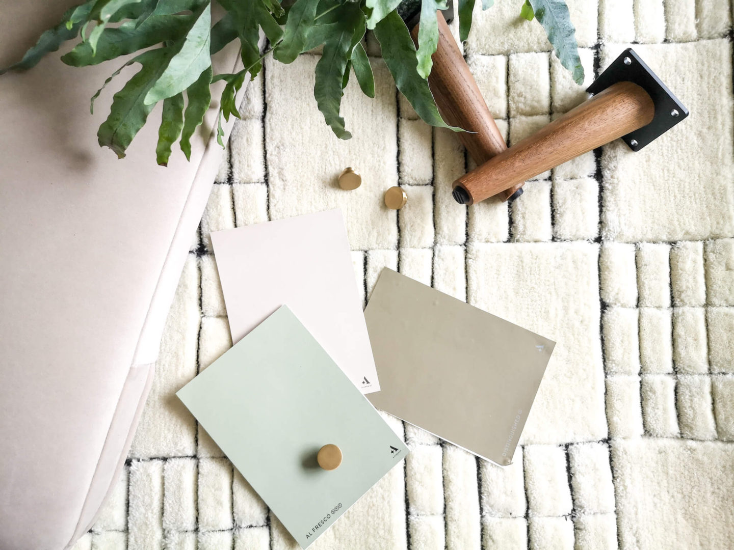 Paint samples next to a gold, round handle, a white and black living room rug, taupe couch cushion and a walnut coffee table leg. 