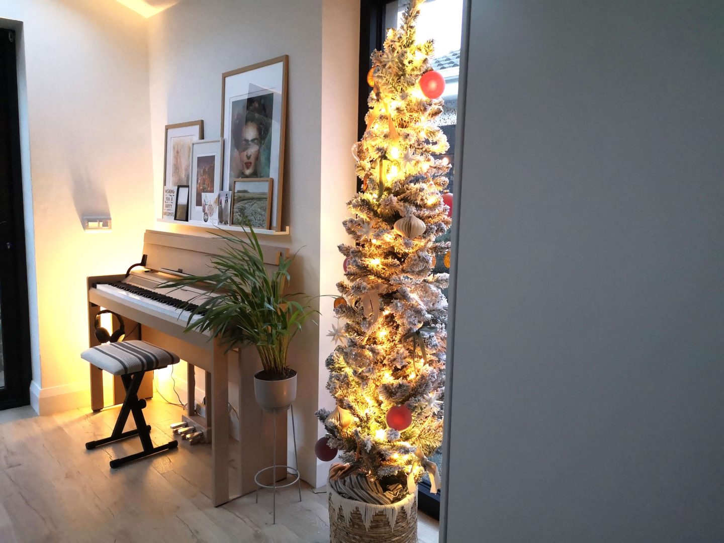 A slim, flocked Christmas tree next to a piano that has a picture above it.