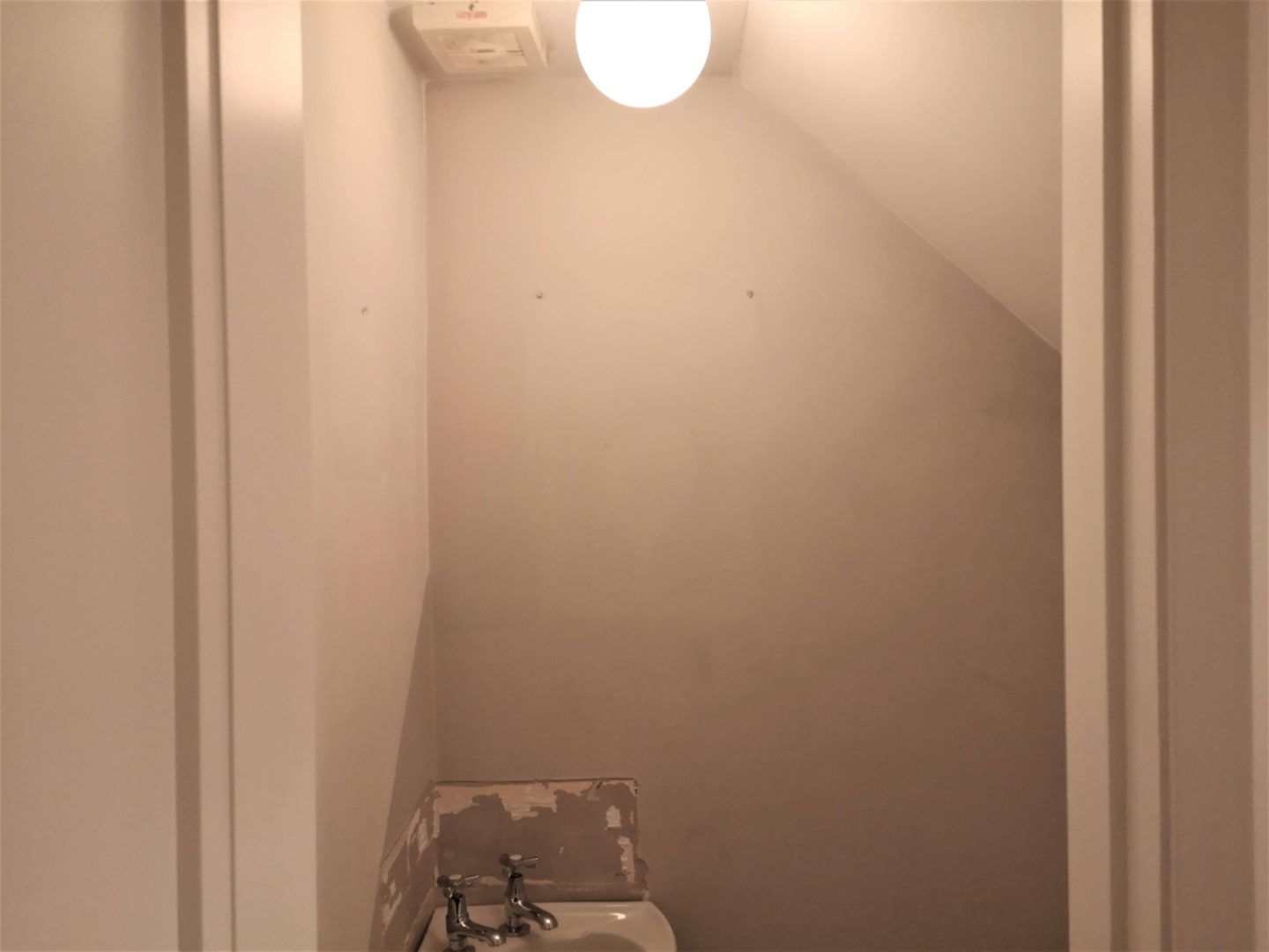 A before picture of a tiny, under the stairs bathroom. It shows plain grey walls and a white sink.